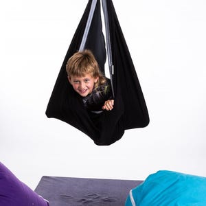 Puede incluir: Un columpio sensorial negro suspendido de un muelle met&aacute;lico. Un ni&ntilde;o est&aacute; dentro del columpio, sonriendo. El columpio cuelga sobre un coj&iacute;n rectangular gris y un puf morado.