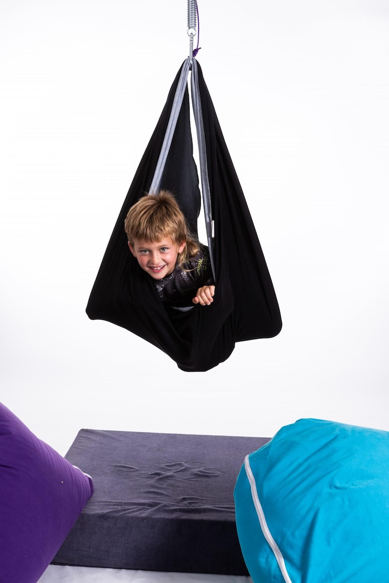 Puede incluir: Un columpio sensorial negro suspendido de un muelle met&aacute;lico. Un ni&ntilde;o est&aacute; dentro del columpio, sonriendo. El columpio cuelga sobre un coj&iacute;n rectangular gris y un puf morado.