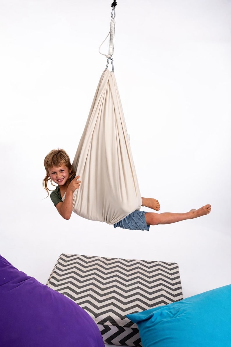 Puede incluir: Un columpio sensorial de tela beige suspendido de un gancho de metal. Un ni&ntilde;o est&aacute; dentro del columpio, sonriendo y mirando a la c&aacute;mara. El ni&ntilde;o lleva una camiseta verde y pantalones cortos azules. Hay pufs en primer plano.
