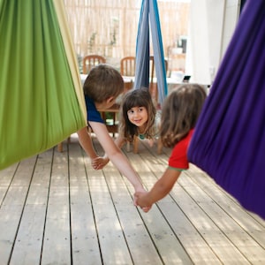 Puede incluir: Ni&ntilde;os jugando en coloridas hamacas colgantes. Las hamacas son en tonos verdes, azules y morados. Los ni&ntilde;os se dan la mano y sonr&iacute;en, disfrutando de la actividad al aire libre en una terraza de madera.