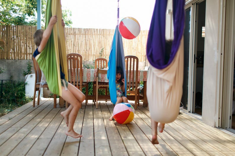 Puede incluir: Ni&ntilde;os jugando al aire libre en coloridas hamacas colgantes. Un bal&oacute;n de playa flota en el aire y otro descansa sobre la cubierta de madera. La escena se desarrolla en un d&iacute;a soleado, con una valla de bamb&uacute; y una mesa y sillas al fondo.