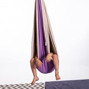 Puede incluir: Un columpio sensorial colgante en beige y morado, suspendido de un resorte y un mosquet&oacute;n. El columpio est&aacute; hecho de tela suave y est&aacute; dise&ntilde;ado para ni&ntilde;os. La imagen muestra a un ni&ntilde;o sentado dentro del columpio, con las piernas colgando.