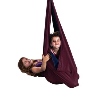 Puede incluir: Un columpio sensorial burdeos con interior azul oscuro, suspendido desde arriba. Dos ni&ntilde;os est&aacute;n dentro del columpio, uno acostado y el otro sentado. Los ni&ntilde;os sonr&iacute;en y miran a la c&aacute;mara.