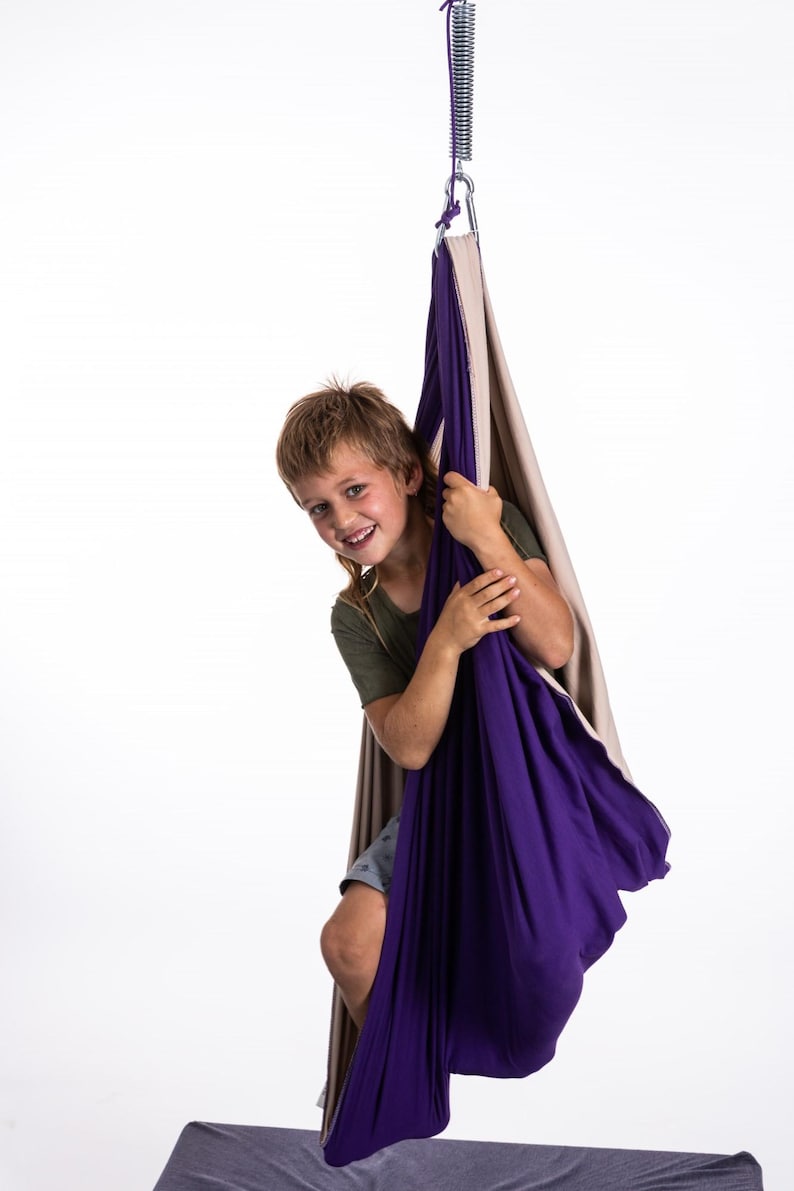 K&ouml;nnte beinhalten: Eine violette und beige sensorische Schaukel, die an einer Metallfeder und einem Haken aufgeh&auml;ngt ist. Die Schaukel besteht aus weichem Stoff und scheint f&uuml;r Kinder konzipiert zu sein. Eine Person ist in der Schaukel und l&auml;chelt.
