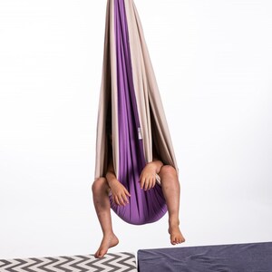 Kid sitting in purple and cream restNest sensory swing chair with spring suspension, indoor hammock swing, Montessori-inspired kids room decor made from soft cotton fabric for sensory regulation and cosy relaxation.