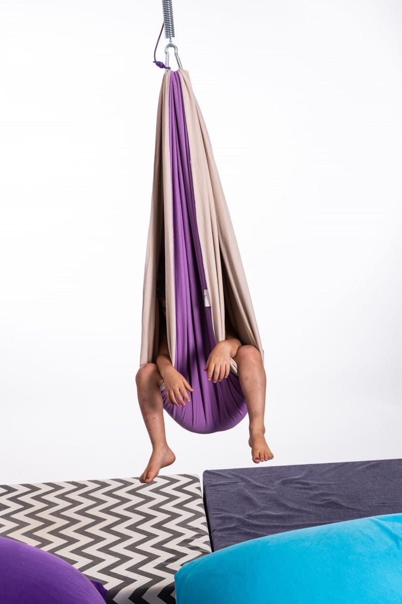 Kid sitting in purple and cream restNest sensory swing chair with spring suspension, indoor hammock swing, Montessori-inspired kids room decor made from soft cotton fabric for sensory regulation and cosy relaxation.