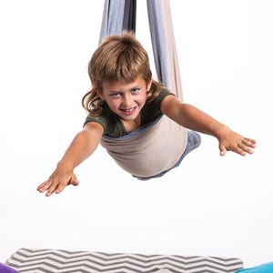 Puede incluir: Un ni&ntilde;o sonr&iacute;e mientras est&aacute; suspendido en una hamaca de tela beige y gris. El ni&ntilde;o lleva una camiseta verde y vaqueros, con los brazos extendidos. La hamaca cuelga desde arriba, con un fondo blanco.