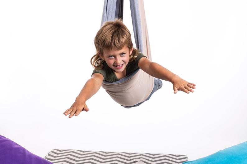 Puede incluir: Un ni&ntilde;o sonr&iacute;e mientras est&aacute; suspendido en una hamaca de tela beige y gris. El ni&ntilde;o lleva una camiseta verde y vaqueros, con los brazos extendidos. La hamaca cuelga desde arriba, con un fondo blanco.