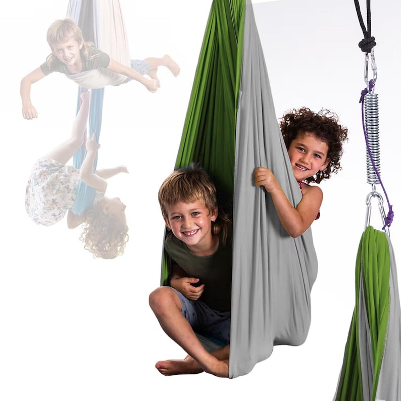 K&ouml;nnte beinhalten: Zwei Kinder l&auml;cheln und spielen in einer gr&uuml;nen und grauen Stoffschaukel. Ein drittes Kind ist im Hintergrund zu sehen, ebenfalls in einer Schaukel. Die Schaukeln sind an der Decke aufgeh&auml;ngt, mit einer Metallfeder und einem Karabiner.