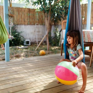 Puede incluir: Dos ni&ntilde;os jugando al aire libre en una terraza de madera. Uno est&aacute; en una hamaca verde y amarilla, y el otro est&aacute; sentado en una hamaca azul y gris, sosteniendo un bal&oacute;n de playa colorido con secciones rosas, verdes y amarillas.