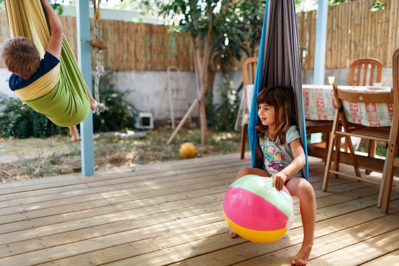 Puede incluir: Dos ni&ntilde;os jugando al aire libre en una terraza de madera. Uno est&aacute; en una hamaca verde y amarilla, y el otro est&aacute; sentado en una hamaca azul y gris, sosteniendo un bal&oacute;n de playa colorido con secciones rosas, verdes y amarillas.