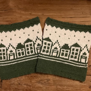 Könnte beinhalten: Zwei gestrickte Weihnachtsstrümpfe mit grün-weißem Design. Die Strümpfe weisen ein sich wiederholendes Muster aus Häusern und kleinen Herzen auf. Die Ober- und Unterseite der Strümpfe sind grün, und jeder hat eine Schlaufe zum Aufhängen.