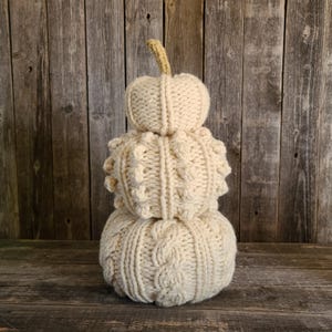 May include: A stacked, cream-coloured knitted pumpkin decoration with a beige stem. The bottom pumpkin has a cable knit design, the middle pumpkin has a textured pattern, and the top pumpkin is smooth. The pumpkins are displayed on a wooden surface.