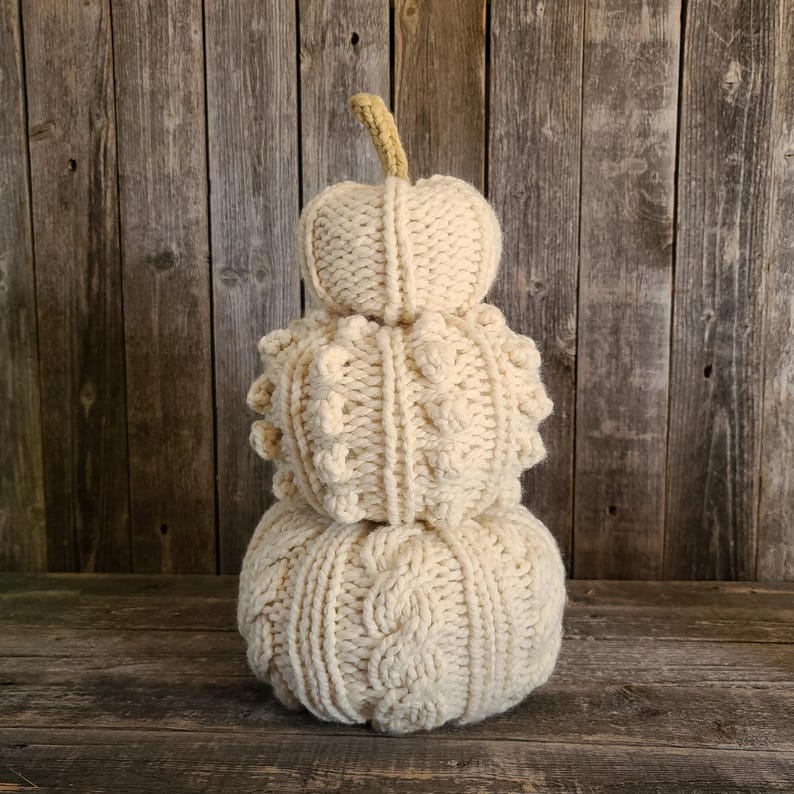 May include: A stacked, cream-coloured knitted pumpkin decoration with a beige stem. The bottom pumpkin has a cable knit design, the middle pumpkin has a textured pattern, and the top pumpkin is smooth. The pumpkins are displayed on a wooden surface.