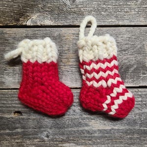 May include: Two miniature, hand-knitted Christmas stockings. One is solid red with a white cuff and hanging loop. The other has a red and white zig-zag pattern on the lower portion. Both are on a weathered wood surface.
