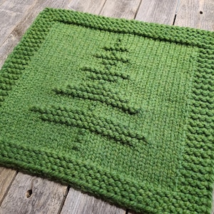 May include: A square, hand-knitted green trivet with a raised Christmas tree design in the center. The trivet has a textured, chunky knit appearance and a thick border. The background is a rustic wooden surface.