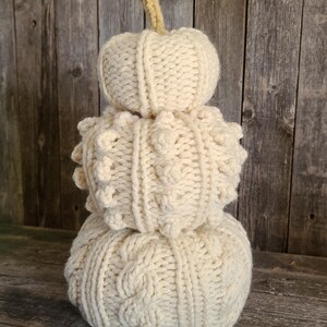 May include: A decorative, off-white, knitted pumpkin. The pumpkin is made of three sections, each with a different texture. The top section is a small, round shape. The middle section has a textured, bumpy pattern. The bottom section has a cable knit design. A beige stem tops the pumpkin.