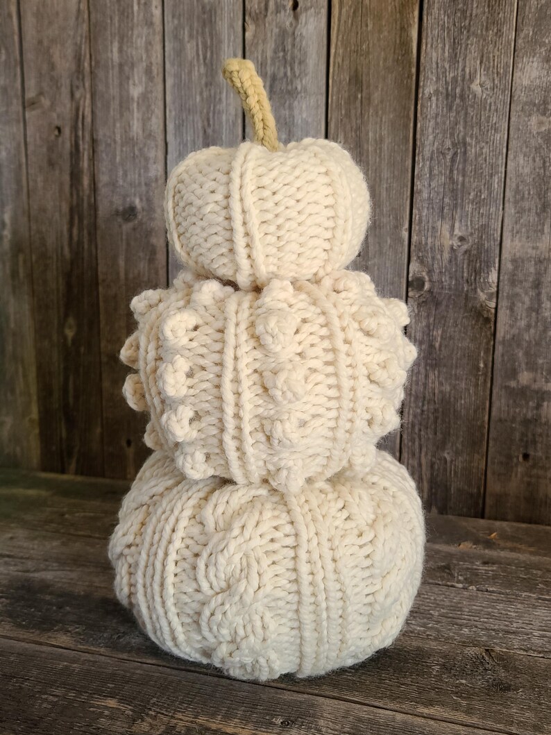 May include: A decorative, off-white, knitted pumpkin. The pumpkin is made of three sections, each with a different texture. The top section is a small, round shape. The middle section has a textured, bumpy pattern. The bottom section has a cable knit design. A beige stem tops the pumpkin.