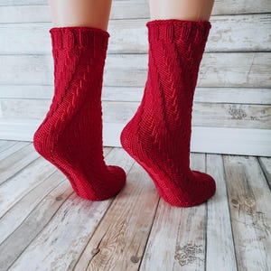 Könnte beinhalten: Ein Paar rote Stricksocken mit einem Zickzackmuster. Die Socken liegen auf einem Holzboden.