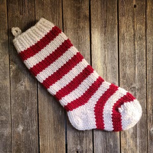 Könnte beinhalten: Ein rot-weiß gestreifter, gestrickter Weihnachtsstrumpf, der an einer Holzwand hängt.