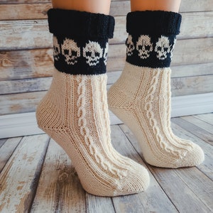 Puede incluir: Un par de calcetines de punto de color crema con diseños de calaveras negras en el puño. Los calcetines están hechos de un material suave y cómodo y tienen un patrón de punto de cable.