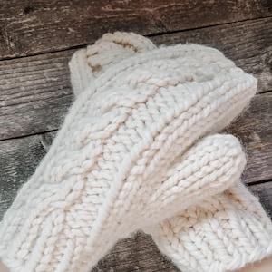 May include: A pair of white knitted mittens with a cable knit pattern. The mittens are made of a soft, fluffy yarn.