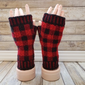 May include: A pair of red and black plaid fingerless gloves. The gloves are made of a knitted material and feature a classic buffalo check pattern. The cuffs are solid black. The gloves are displayed on mannequin hands.