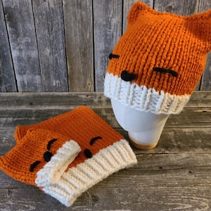 May include: Two knitted fox hats with orange and white stripes and black embroidered eyes. The hats have floppy ears.