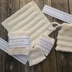 May include: A set of four white knitted kitchen items: a dishcloth, two scrubbies, and a soap saver. Each item has a label with the words "Handknit & Reusable" and the name of the item.