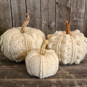 May include: Three cream-coloured, knitted pumpkins of varying sizes. The pumpkins have textured cable knit patterns and are topped with natural stems. They are arranged on a rustic wooden surface.