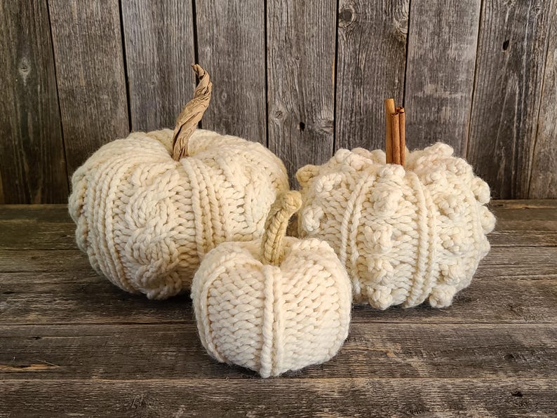May include: Three cream-coloured, knitted pumpkins of varying sizes. The pumpkins have textured cable knit patterns and are topped with natural stems. They are arranged on a rustic wooden surface.
