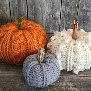 May include: Three knitted pumpkins in orange, cream, and grey, with textured surfaces and stems. The orange pumpkin has a cable knit design, the cream pumpkin has a textured bobble stitch, and the grey pumpkin is smaller. They are on a rustic wooden surface.
