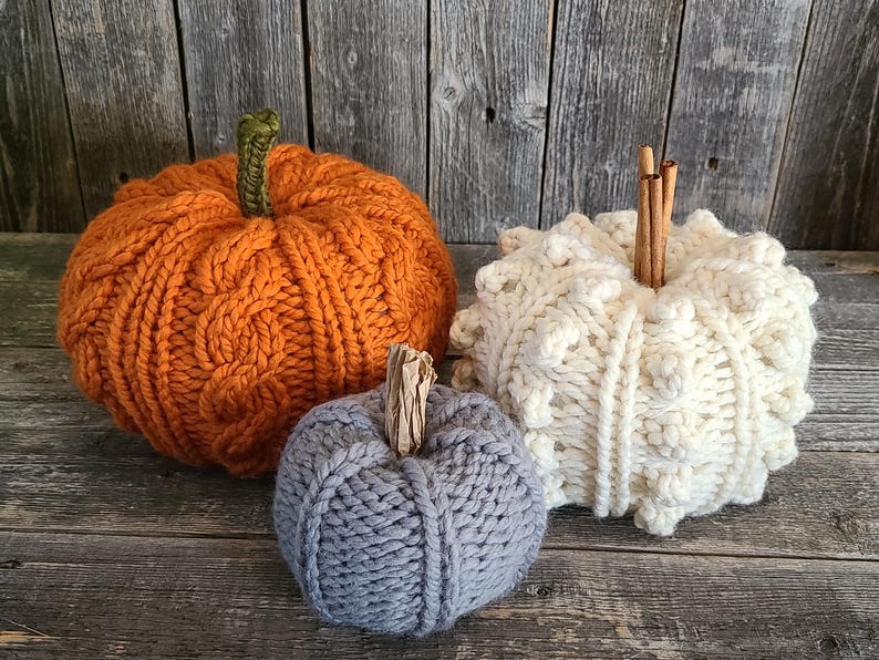 May include: Three knitted pumpkins in orange, cream, and grey, with textured surfaces and stems. The orange pumpkin has a cable knit design, the cream pumpkin has a textured bobble stitch, and the grey pumpkin is smaller. They are on a rustic wooden surface.