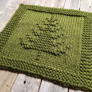 May include: A square, knitted item in shades of green, featuring a textured tree design in the center. The edges have a thicker border. The item is placed on a rustic wooden surface.