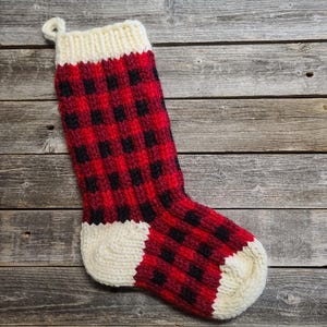 May include: A hand-knitted Christmas stocking with a red and black buffalo plaid pattern. The cuff and toe are cream-coloured. A loop is attached for hanging. The stocking is displayed on a weathered wooden surface.