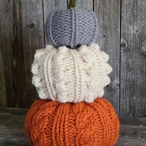 May include: A decorative stack of three knitted pumpkins. The base pumpkin is orange with a cable knit pattern, the middle is cream with a textured design, and the top is grey with a simple knit. A green stem sits atop the grey pumpkin. The pumpkins are for autumn decor.