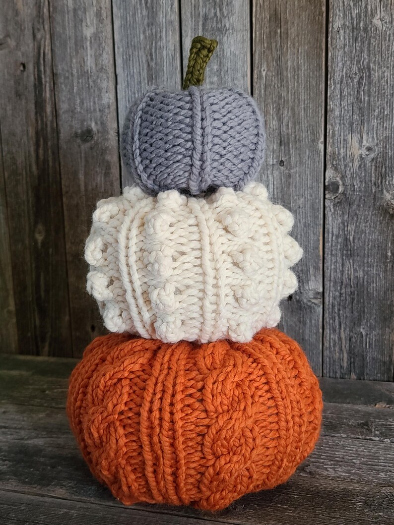 May include: A decorative stack of three knitted pumpkins. The base pumpkin is orange with a cable knit pattern, the middle is cream with a textured design, and the top is grey with a simple knit. A green stem sits atop the grey pumpkin. The pumpkins are for autumn decor.