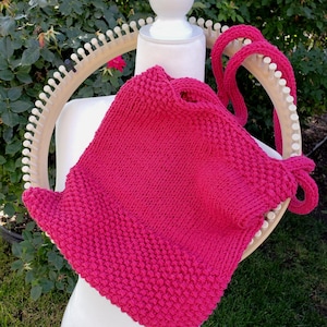 Peut inclure: Un sac tricoté rose vif avec des sangles assorties est présenté sur un mannequin blanc. Le sac est fait d'un point texturé et possède une fermeture à cordon. Un métier à tricoter en bois est derrière le sac.
