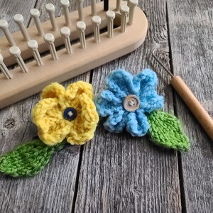 May include: A wooden knitting loom and crochet hook sit beside two handmade crochet flowers. One flower is yellow with a blue button center, the other is blue with a wooden button center. Both have green leaves.