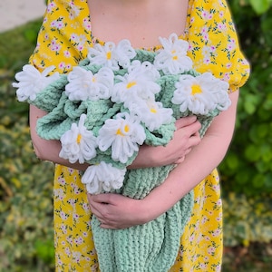 May include: A crochet blanket in a soft sage green colour featuring multiple three-dimensional white daisy flowers with bright yellow centres attached to the surface. The blanket is made of a thick, plush yarn with a visible looped texture.
