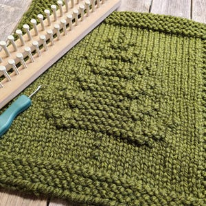 Puede incluir: Un cuadrado tejido a mano de color verde con un diseño de árbol, un telar de punto de madera y un ganchillo turquesa. La pieza tejida mide aproximadamente 30 cm cuadrados, con un patrón de árbol texturizado en el centro.