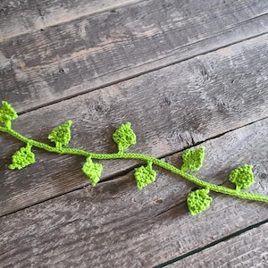 May include: A green crocheted vine with leaves, on a wooden background.