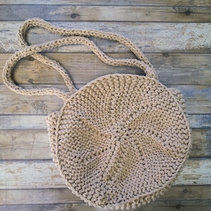 Könnte beinhalten: Eine beige runde Tasche aus Häkelarbeit mit einem langen Riemen. Die Tasche ist aus dickem Garn gefertigt und hat eine strukturierte Oberfläche.