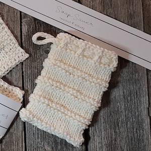 May include: White knitted soap saver bag with the text "Soap Saver Handknit & Reusable" on a white tag. The bag is on a wooden surface.