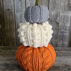 Op de afbeelding: Een stapel van drie gebreide pompoenen in oranje, crème en grijs. De onderste pompoen is oranje met een kabelgebreid patroon. De middelste pompoen is crèmekleurig met een textuurpatroon. De bovenste pompoen is grijs met verticale ribbels en een groene steel.