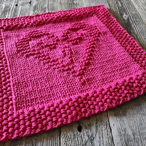 May include: A pink knitted dishcloth with a heart design. The dishcloth is made with a textured stitch that gives it a raised appearance.
