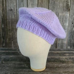 Op de afbeelding: Een lichtpaarse gebreide baret. De baret wordt getoond op een mannequinhoofd. De hoed is gemaakt van een zacht, getextureerd garen en heeft een geribbelde rand. De baret is een klassieke stijl, perfect om een vleugje elegantie aan elke outfit toe te voegen.