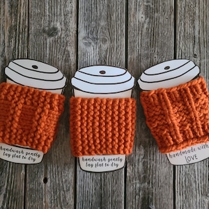 Puede incluir: Tres calientavasos de punto naranja con insertos de papel blanco para vasos de café. Los calientavasos son hechos a mano y tienen instrucciones para lavarse suavemente a mano y dejarse secar en plano. Un calientavasos tiene el texto "hecho a mano con AMOR" en el inserto de papel.