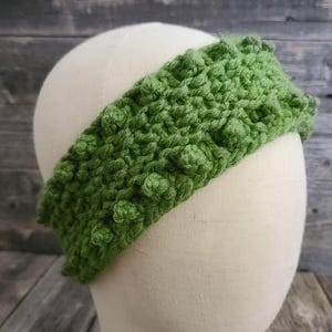 May include: A green, textured crochet headband displayed on a mannequin head. The headband is a vibrant shade of green and features a raised, bobble-like stitch pattern. The background is a rustic wooden surface.