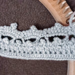 May include: A close-up of a gray crocheted crown being worked on a wooden loom. The crown has a black design and is being worked with a light gray yarn.